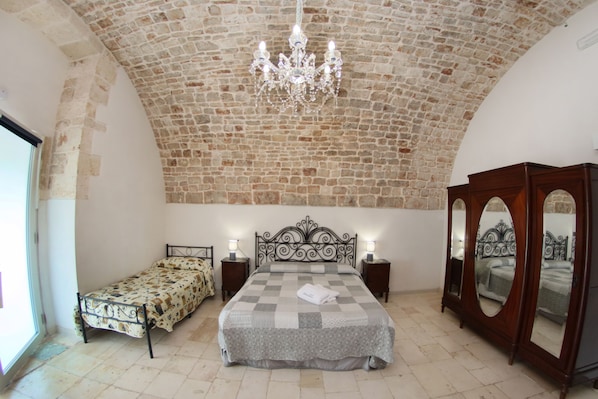 A bedroom with a stone ceiling, a chandelier, a bed with a checkered bedspread, two bedside tables with lamps, and a large mirror. - Masseria Alberotanza (Conversano)