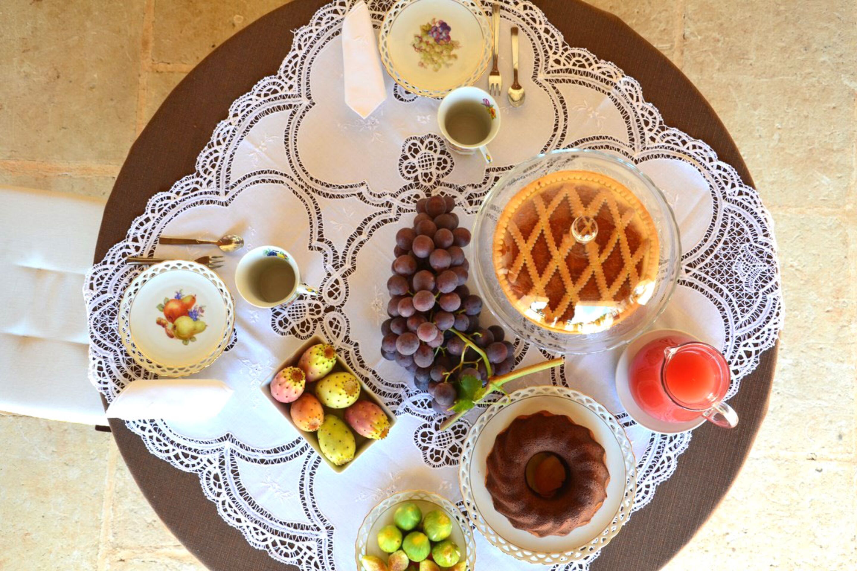 A table set with a lace tablecloth, featuring a plate of grapes, a glass of red juice, a chocolate dessert, a bowl of fruit, and a cup of tea.