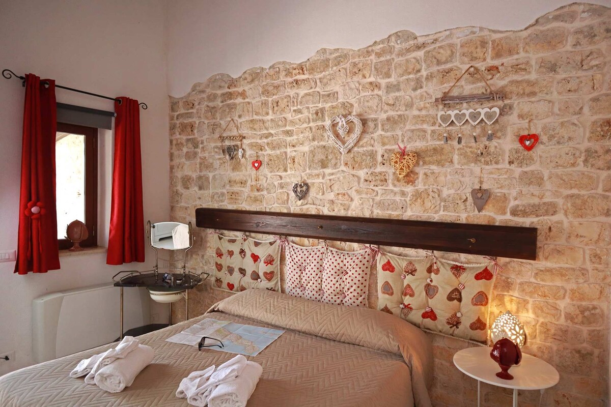 Un dormitorio con pared de piedra, cabecero de madera, mesita de noche con lámpara y ventana con cortinas rojas.