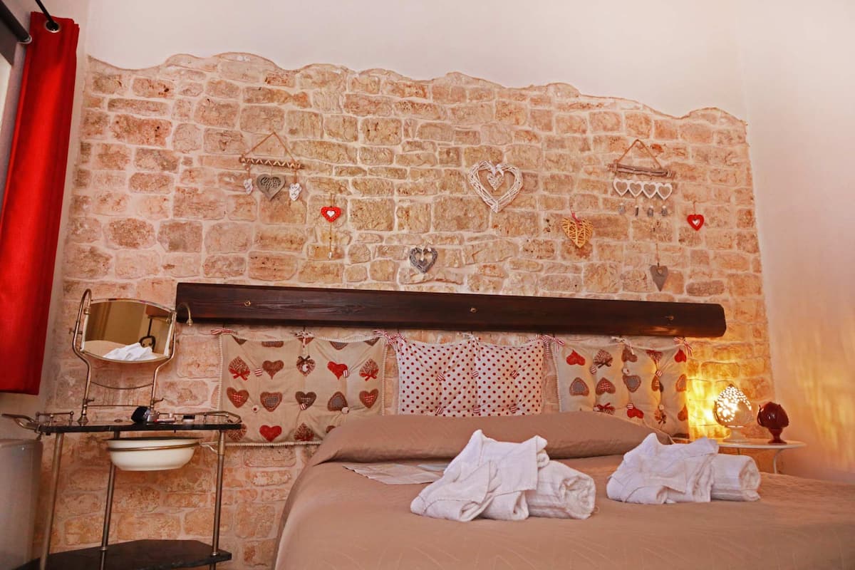 A bedroom with a stone wall, a bed with white linens, a bedside table with a mirror, and heart-shaped decorations.