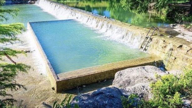 Une piscine rectangulaire bordée de béton, avec un escalier menant à l’eau, entourée de rochers et de verdure.