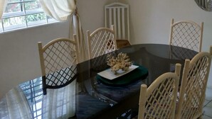 Un comedor con una mesa de vidrio rodeada de sillas de mimbre, un adorno de coral y una ventana con cortinas.