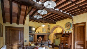 Ein rustikaler Essbereich mit Holztisch, gedecktem Tisch für eine Mahlzeit, Kamin und Küchenzeile mit Holzschränken und einer Uhr an der Wand.