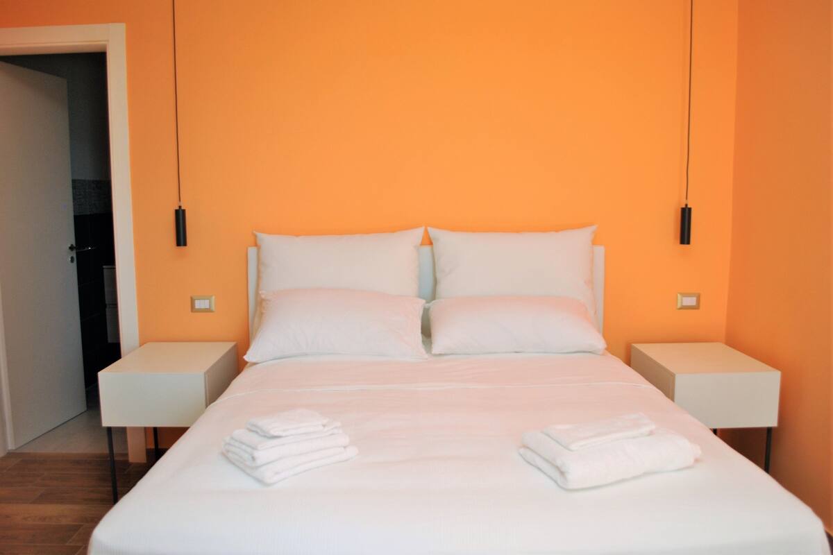 Une chambre d’hôtel avec un grand lit, du linge de lit blanc, deux tables de chevet et un mur orange.