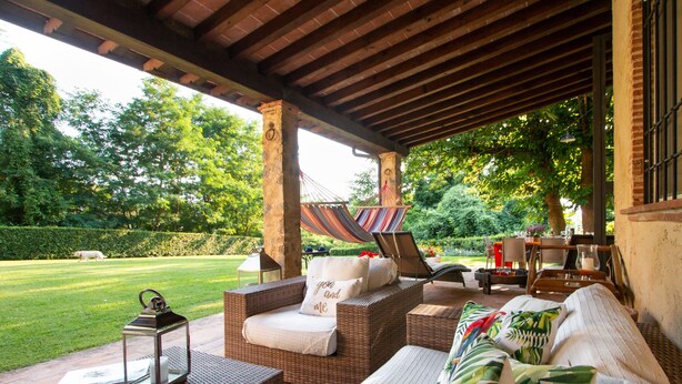 A covered outdoor seating area with wicker furniture, a hammock, and a view of a green lawn and trees. - Il Castagno (Camaiore)
