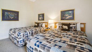 A bedroom with two beds, each with a checkered blanket and bear-themed pillows. There are framed paintings on the walls and a bedside lamp on one bed.