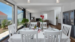 A modern dining area with a table set for four, white chairs, and a vase of red flowers. A sofa and a television are visible in the background. - Modern house with private pool, family-friendly, WiFi, barbecue, sea view, quiet (Makarska)