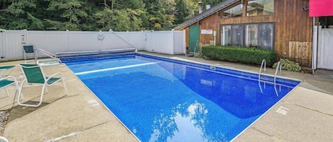 Une piscine rectangulaire à l’eau d’un bleu limpide, entourée d’une terrasse en béton. On y trouve des chaises longues vertes et un petit bâtiment en bois doté d’une fenêtre.