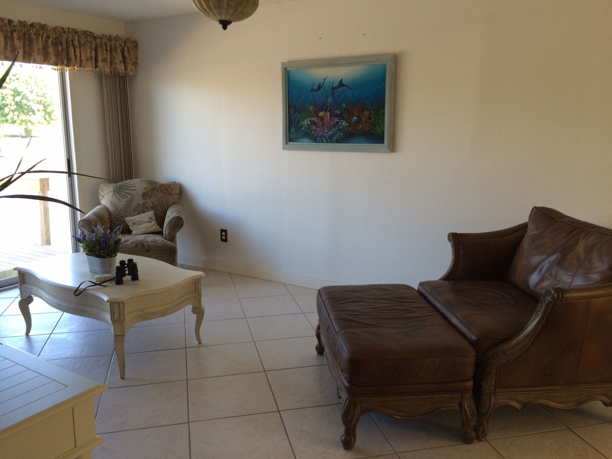 A living room with a brown leather sofa, a white armchair, a small table with binoculars, and a painting of underwater scenery.
