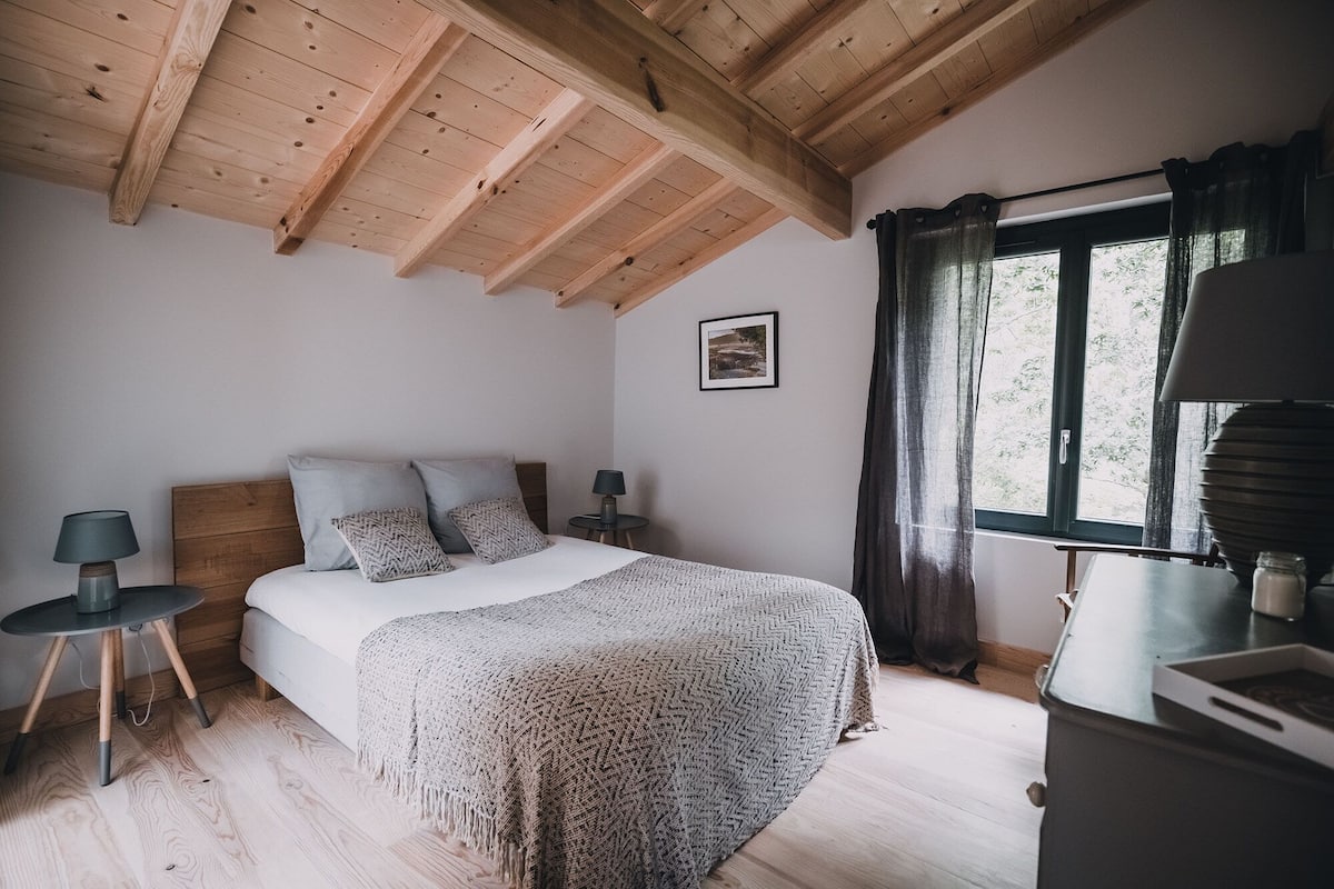 A bedroom with a wooden ceiling, a bed with a patterned blanket, two bedside tables with lamps, a window with curtains, and a framed picture on the wall.