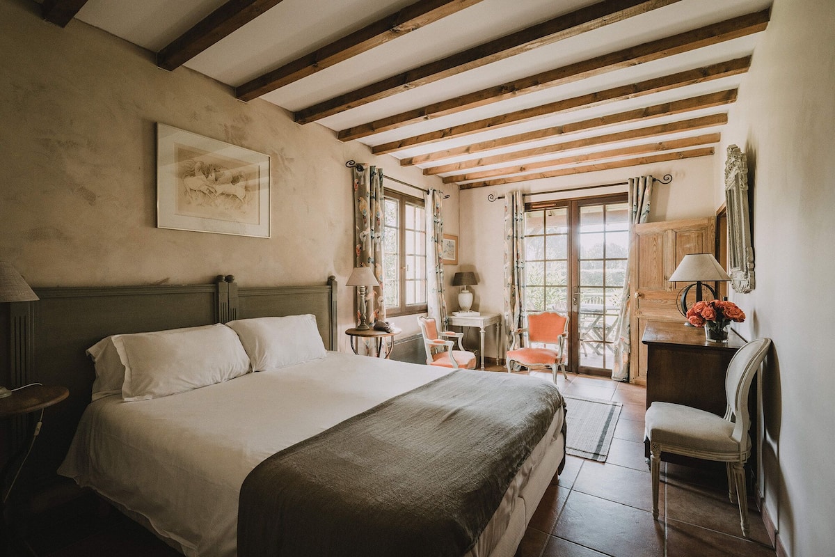 A bedroom with a large bed, wooden beams on the ceiling, a desk with a lamp, and a chair.
