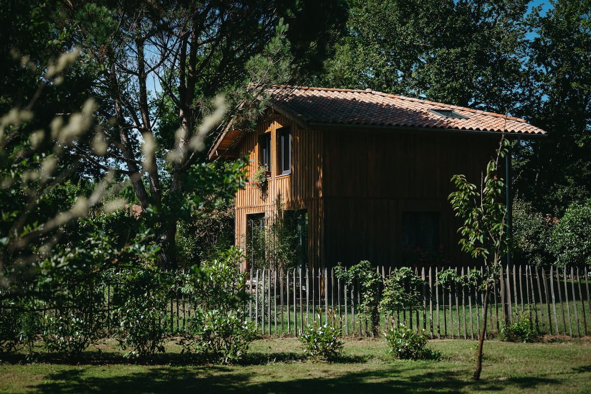 A wooden cabin with a tiled roof, surrounded by a wooden fence and lush greenery.