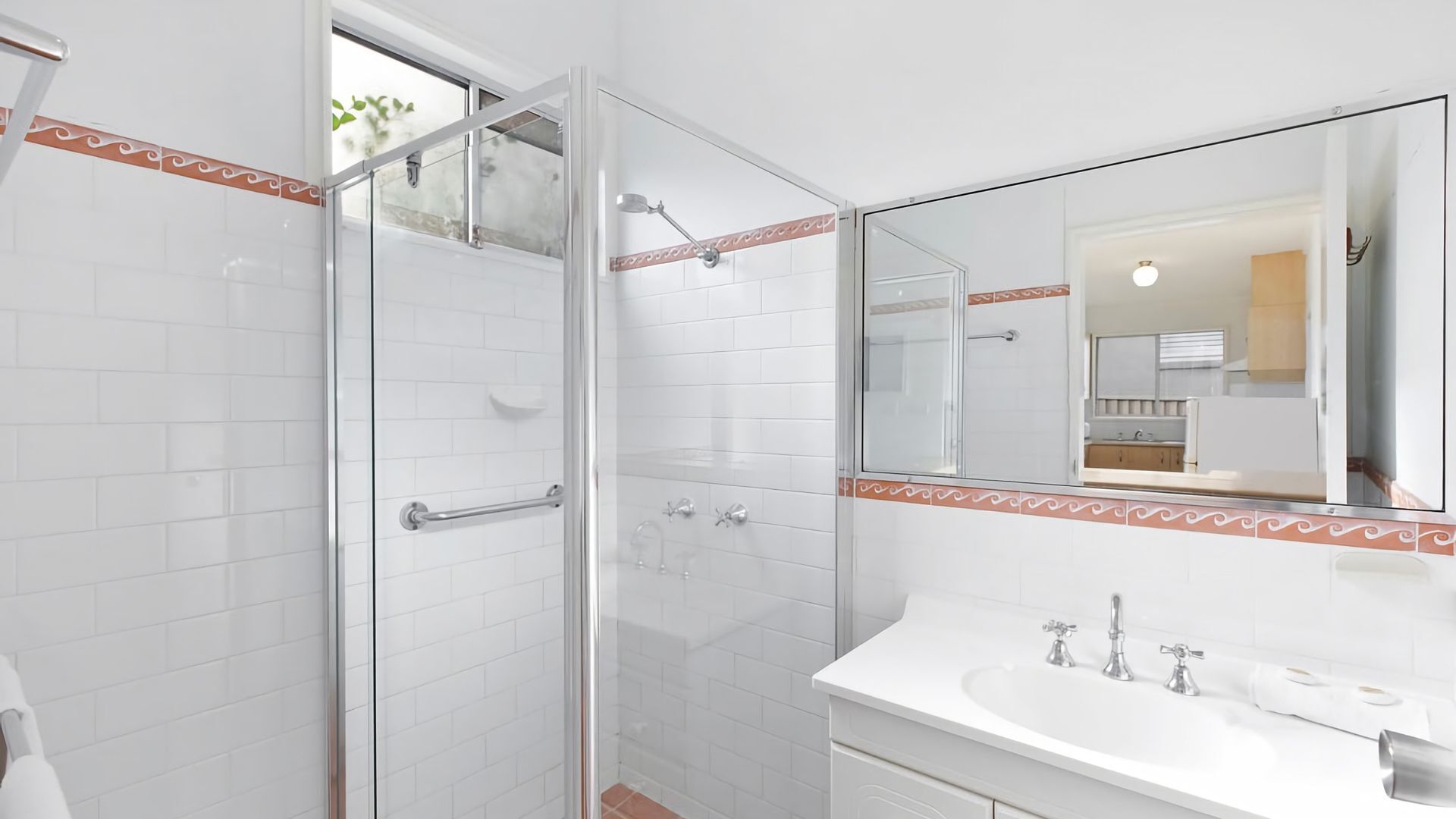 A modern bathroom with a glass shower enclosure, white tiled walls, a white sink with a mirror above it, and a decorative border around the mirror.