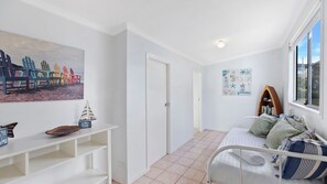 Une pièce avec un lit, un mur blanc, une porte, une étagère sur laquelle est posé un modèle de bateau et un tableau représentant des chaises de plage.