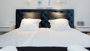Een netjes opgemaakt bed met witte beddengoed en twee kussens, een donkerblauwe hoofdbord en twee wandlampen.