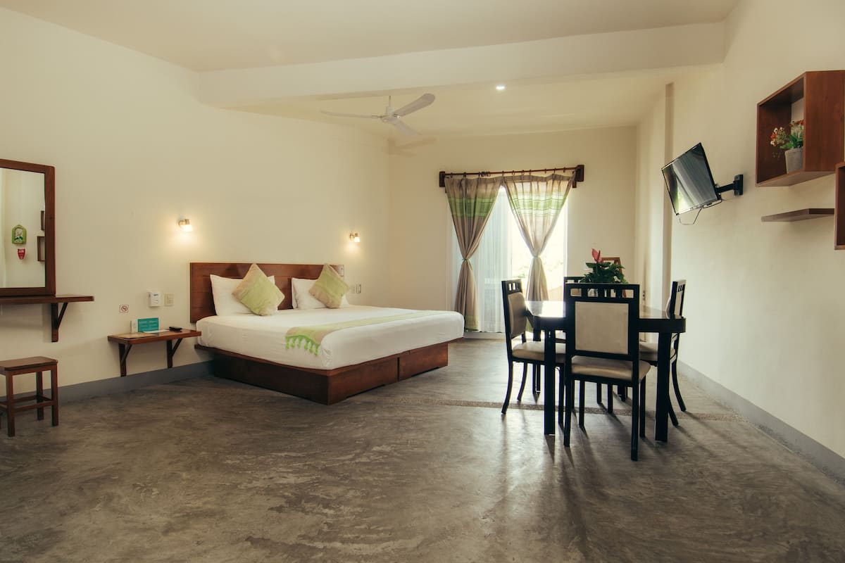 A bedroom with a bed, bedside tables, a desk with chairs, a ceiling fan, and a TV mounted on the wall.