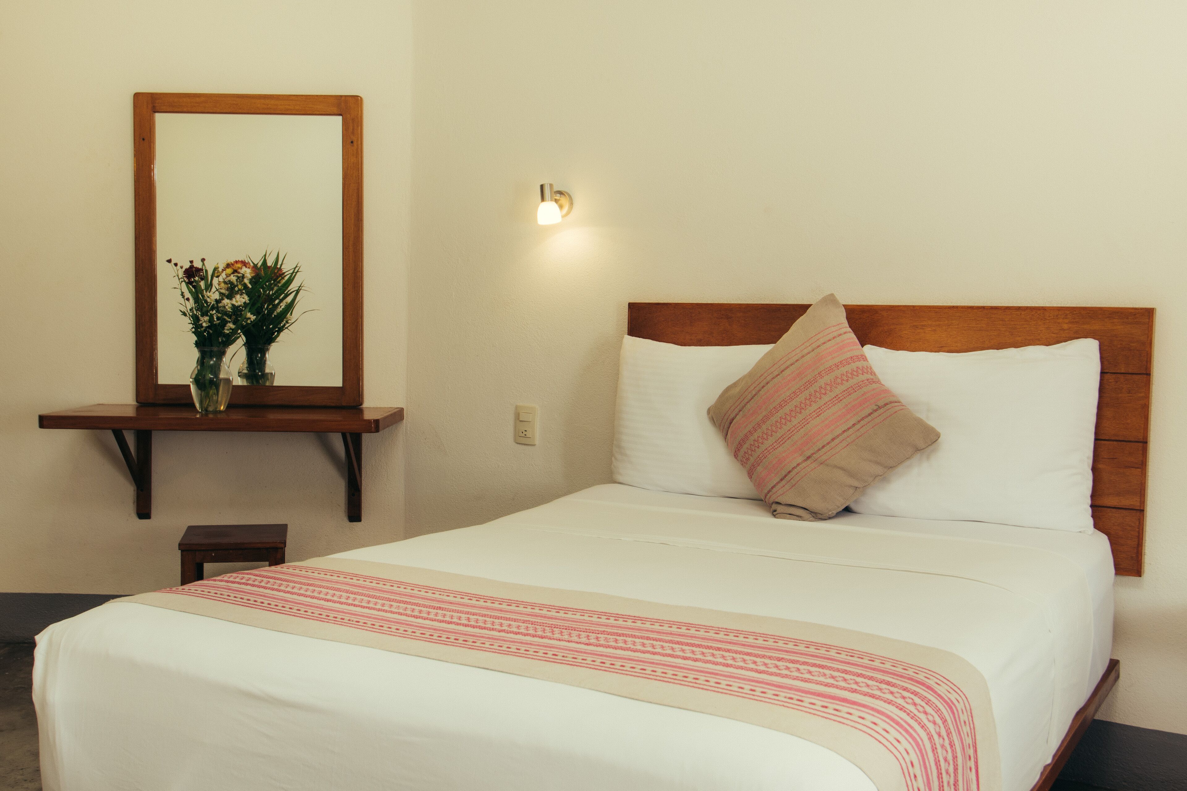 Een net opgemaakt bed met een witte deken en een roze-wit gestreepte plaid. Er is een houten hoofdbord, een nachtkastje met een bloevas en een wandlamp.