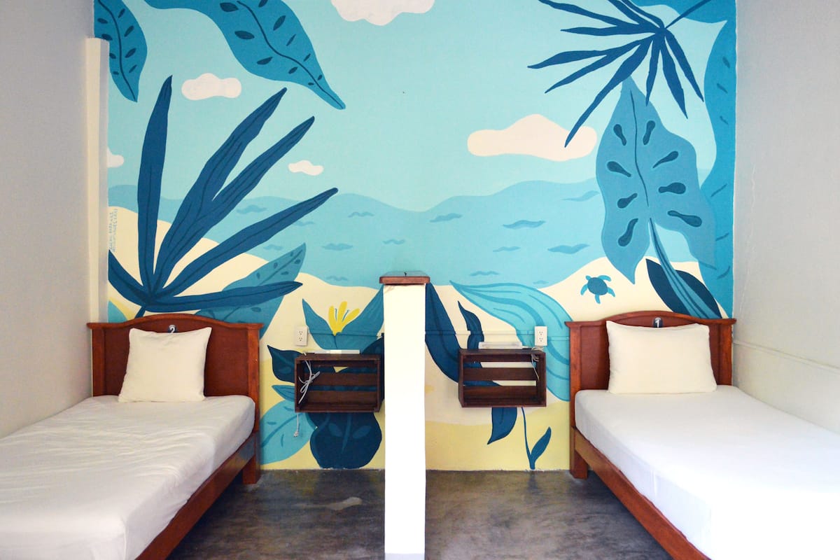 A room with two single beds, a wooden headboard, a wall mural with tropical leaves, and a small shelf.