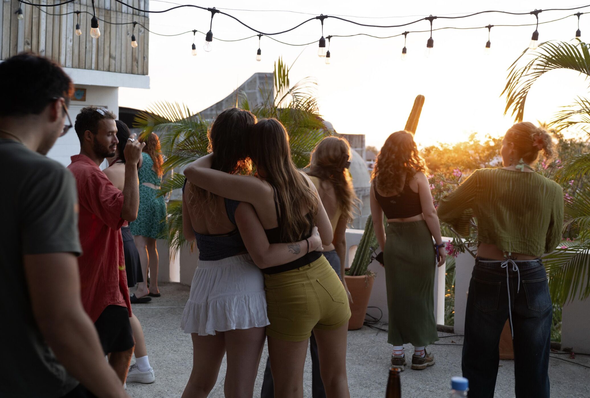 A group of people outdoors, possibly at a social gathering, with string lights overhead and palm trees in the background.