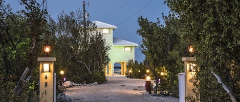 A house with a gravel driveway leading to a gated entrance, surrounded by trees and a clear sky.