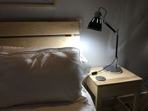Una cama con ropa blanca, cabecera de madera, mesita de noche con lámpara y un enchufe de pared.