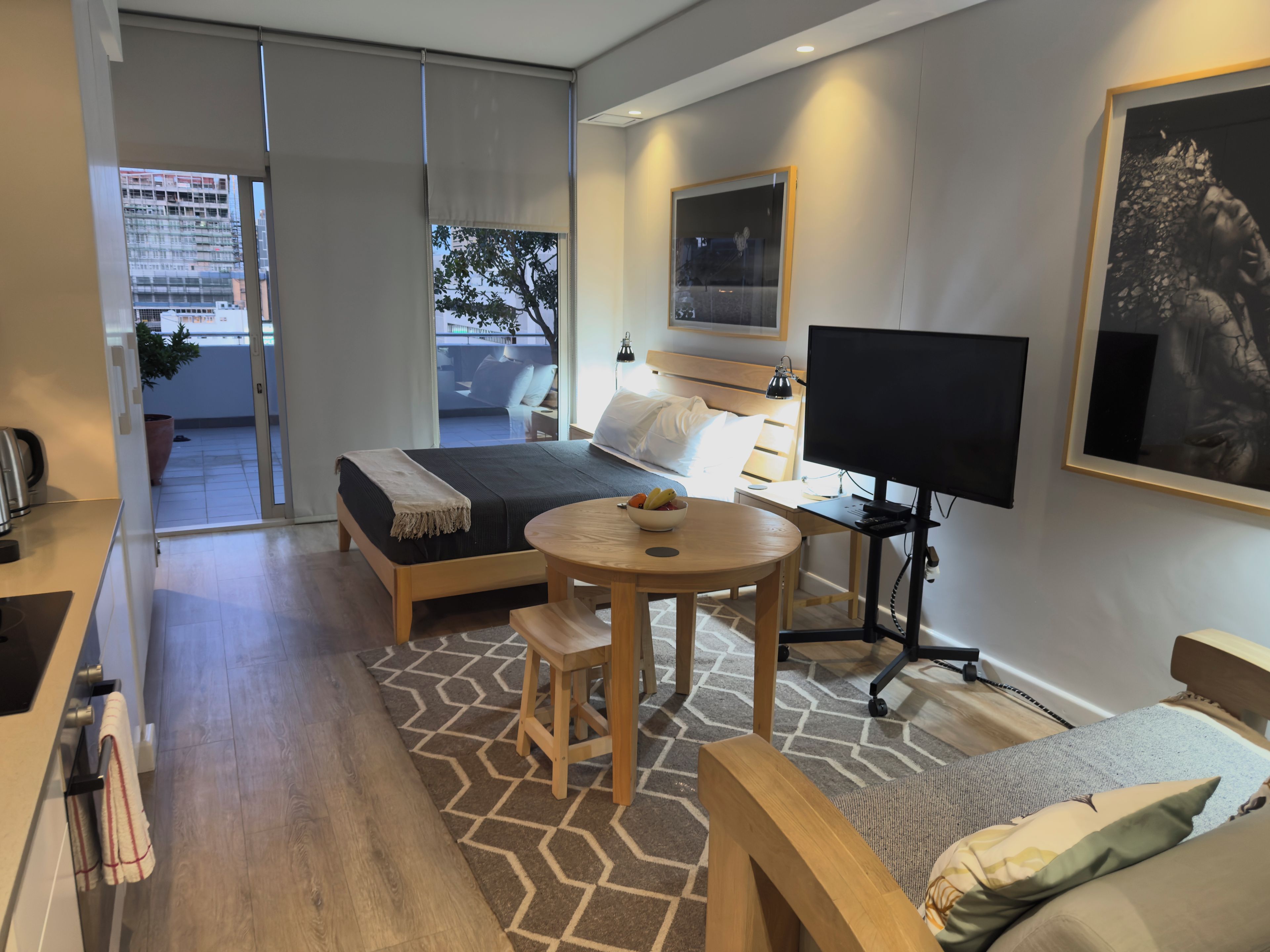 A modern hotel room with a bed, a desk with a computer, a chair, a small table with fruit, and a television.