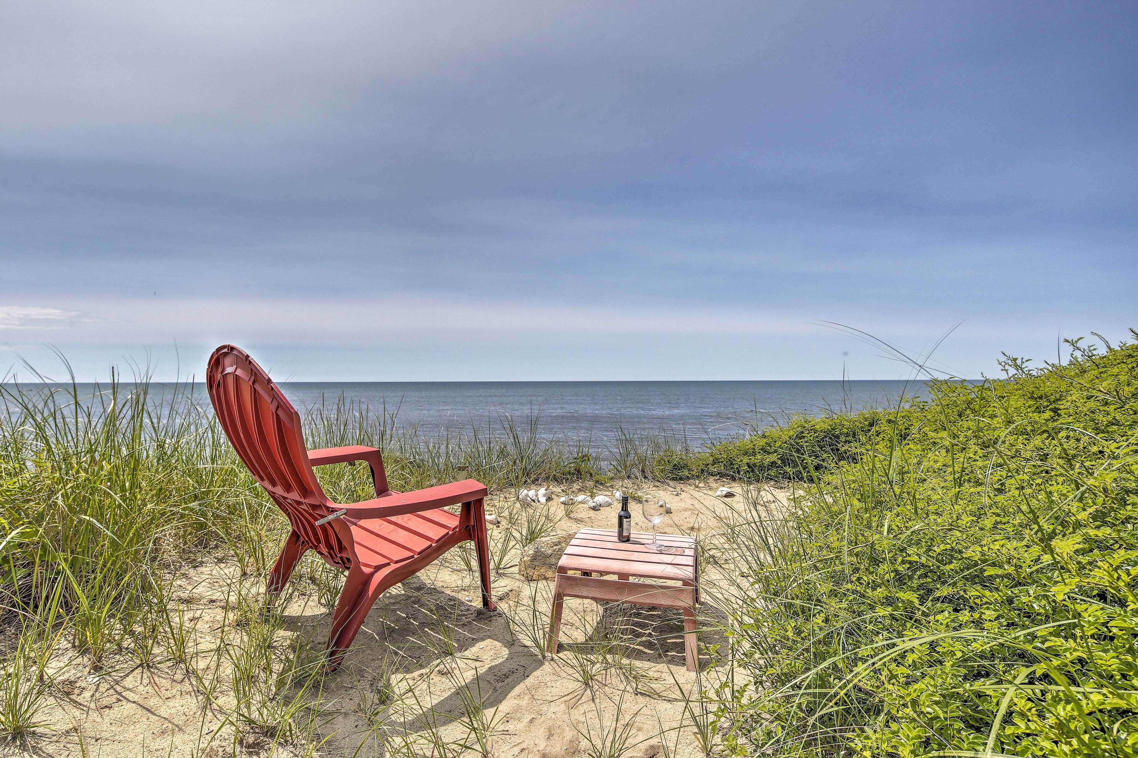 A red Adirondack chair and a small wooden table with a bottle and glass on a sandy beach with grass and the ocean in the background.