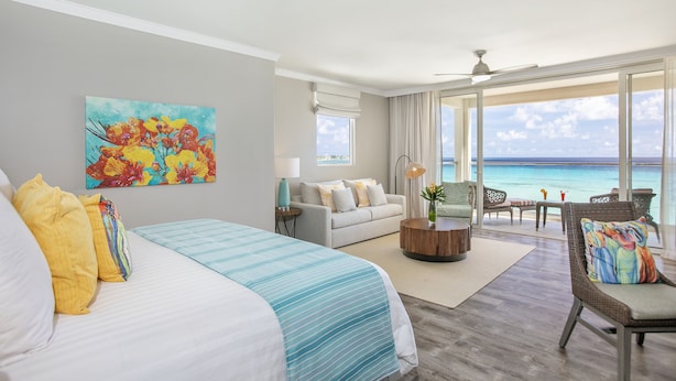 Habitación de hotel con cama, sofá, zona de comedor y un amplio ventanal con vista al océano.