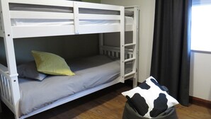 Una cama litera con colchón gris y almohada verde, un sillón de peluche blanco y negro, y una ventana con cortinas oscuras.