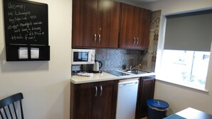 Una cocina compacta con armarios de madera oscura, un microondas blanco, una cafetera y un lavabo. Hay una pizarra con un mensaje de bienvenida y detalles de Wi-Fi. Se ve una silla y una mesa en primer plano.