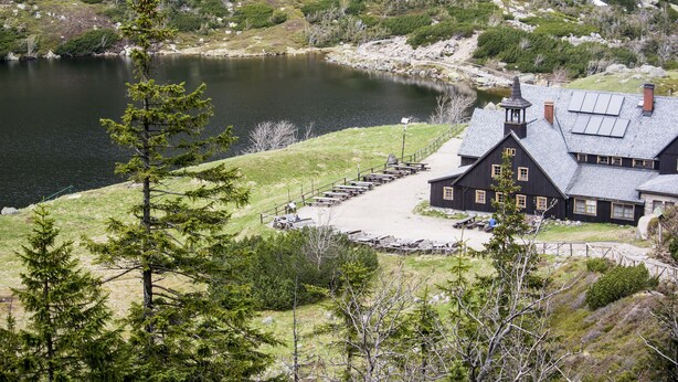 Una cabaña con una chimenea destacada, un muelle que se adentra en un lago y un paisaje boscoso circundante.