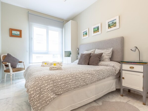 A bedroom with a bed, a chair, a nightstand, and a window.