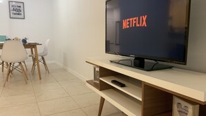 A living room with a flat-screen TV displaying 'NETFLIX' on the screen, a dining area with a table and chairs, and a framed picture on the wall.