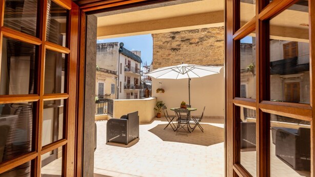 Un balcón abierto con una mesa y sillas, una sombrilla blanca y vistas a edificios al fondo.