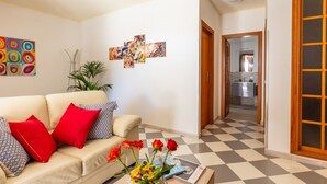 Una sala de estar con un sofá color beige, cojines rojos y con estampado, una mesa de centro con flores y un suelo a cuadros. En la pared hay pinturas abstractas y una puerta de madera que da a otra habitación.