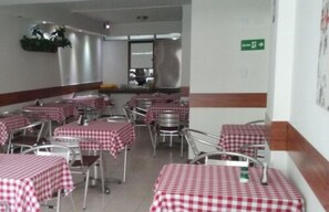 A dining area with multiple tables covered in red and white checkered tablecloths, metal chairs, and a kitchen area in the background. - Hotel San Fermin Pereira (Pereira)