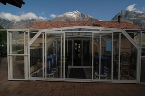Une structure entièrement vitrée, surmontée d’un toit recouvert de tuiles rouges, dotée d’une porte et d’un petit espace extérieur aménagé avec des tables et des chaises.