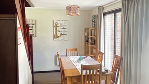A dining area with a wooden table and chairs, a hanging light fixture, a bookshelf, and a window with curtains.