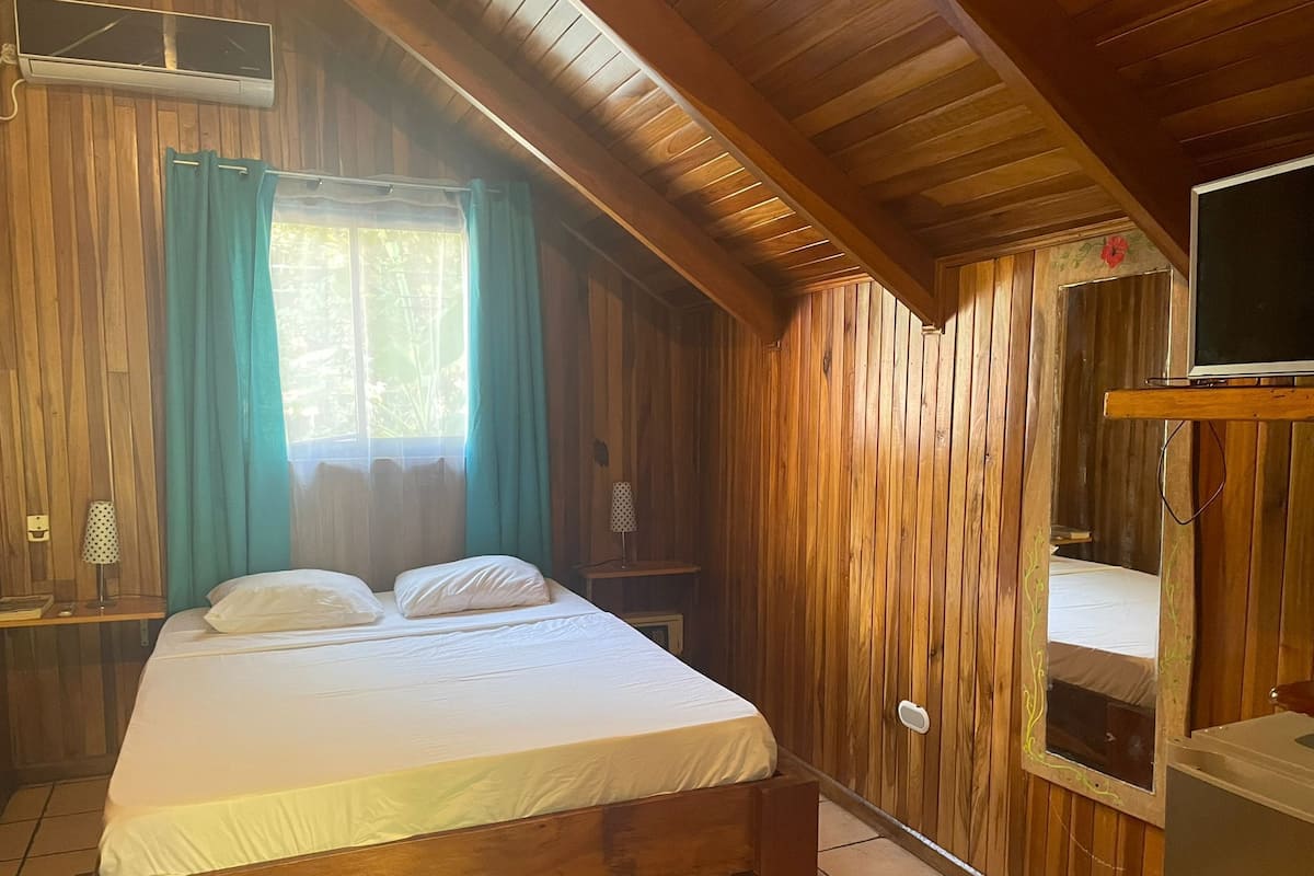 A wooden room with a bed, a window with curtains, an air conditioning unit, a television, and a small table with a lamp.