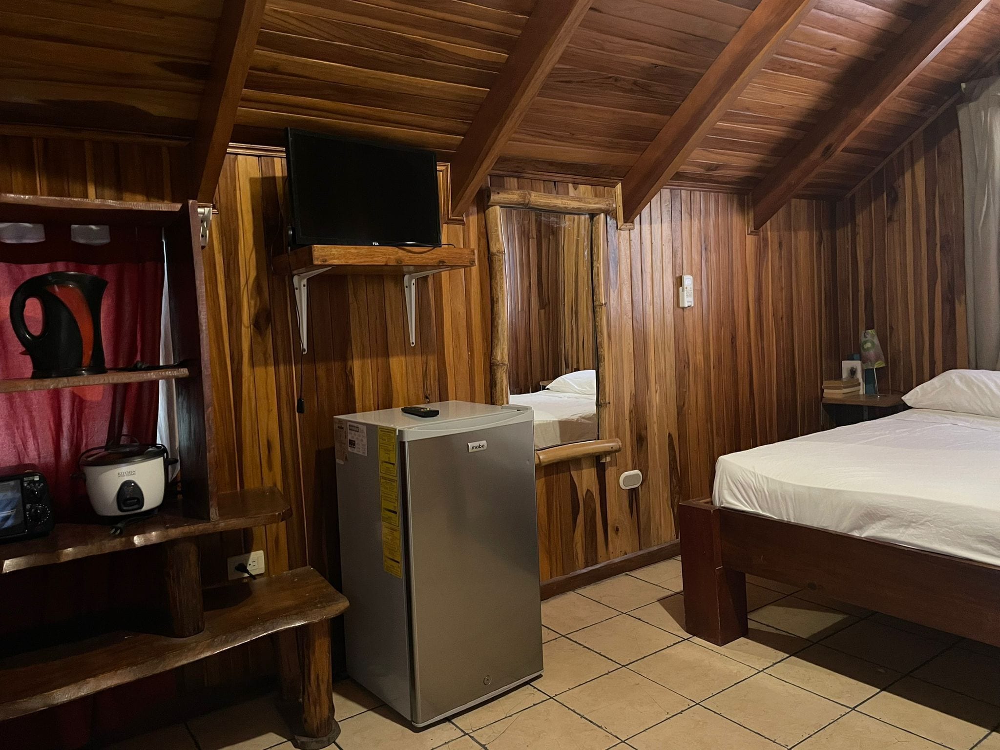 A compact room with wooden walls, a bed, a small refrigerator, a television mounted on the wall, and a shelf with kitchen items.