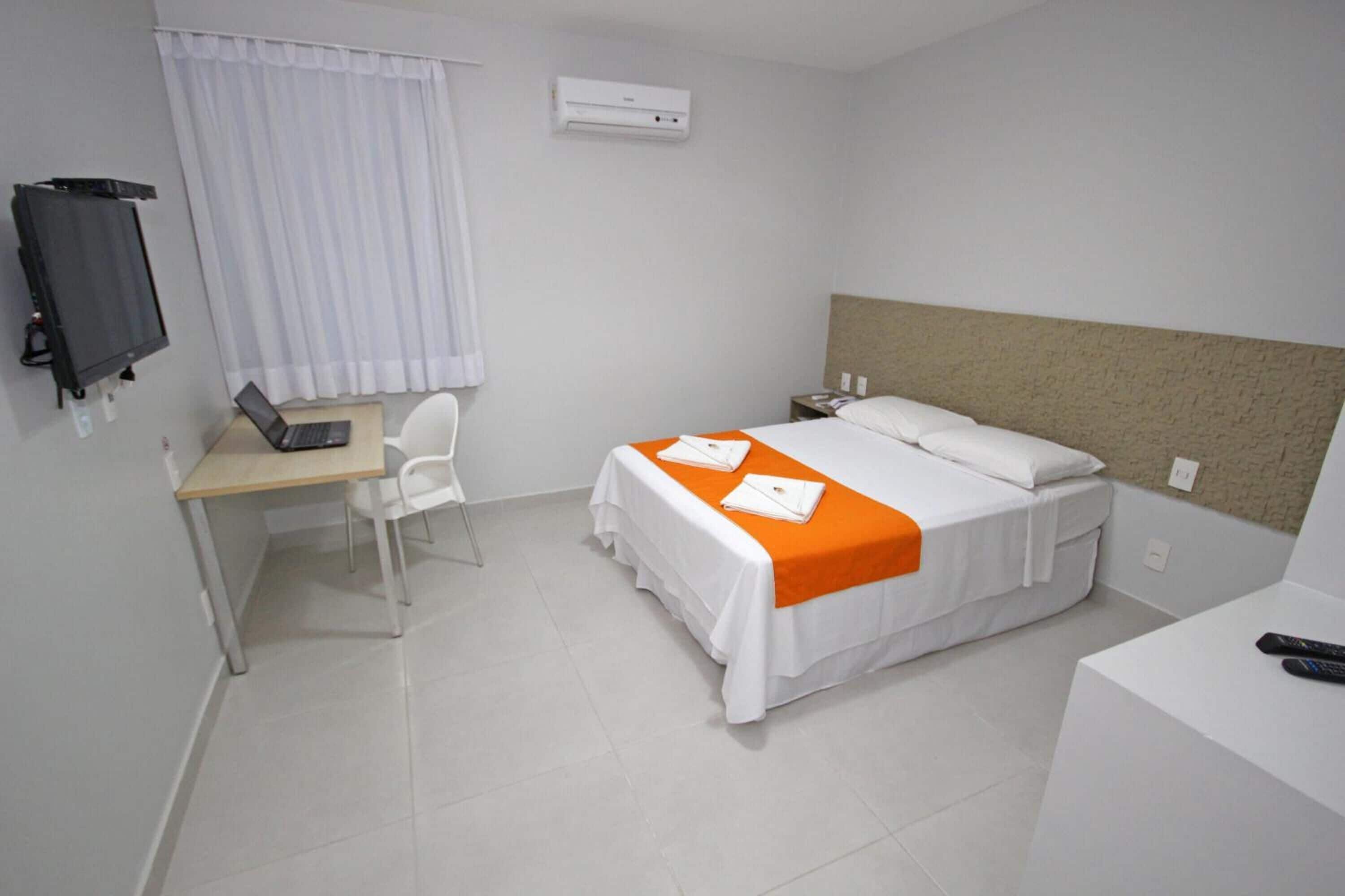 Quarto de hotel com uma cama, uma escrivaninha com um laptop, uma cadeira, uma televisão e uma mesa de cabeceira com controle remoto.