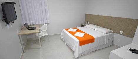 A hotel room with a bed, a desk with a laptop, a chair, a TV, and a bedside table with a remote control.