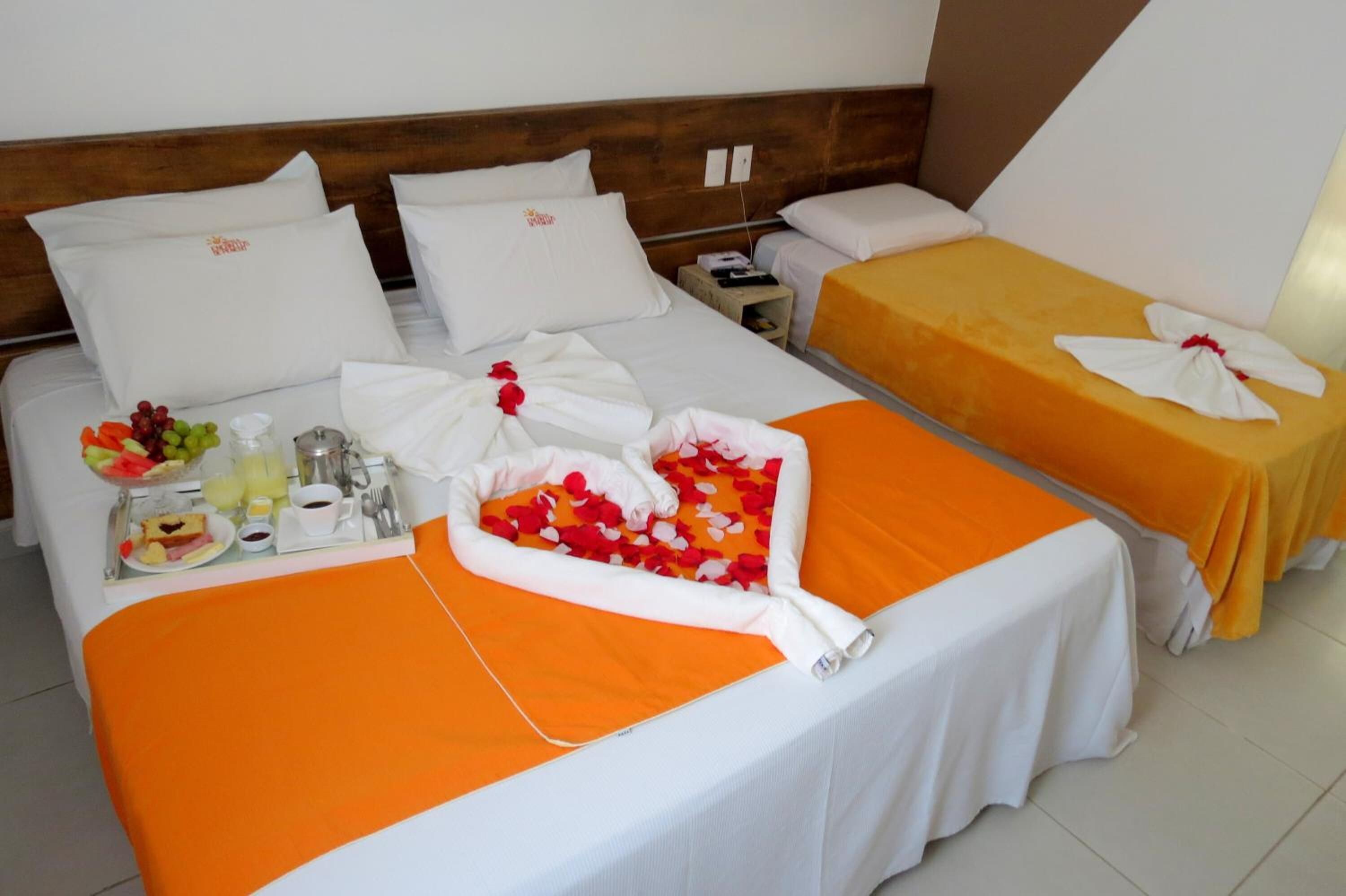 Quarto de hotel com duas camas, cada uma com colcha laranja e almofadas brancas. Há uma bandeja com itens de café da manhã, incluindo frutas, suco e uma xícara.