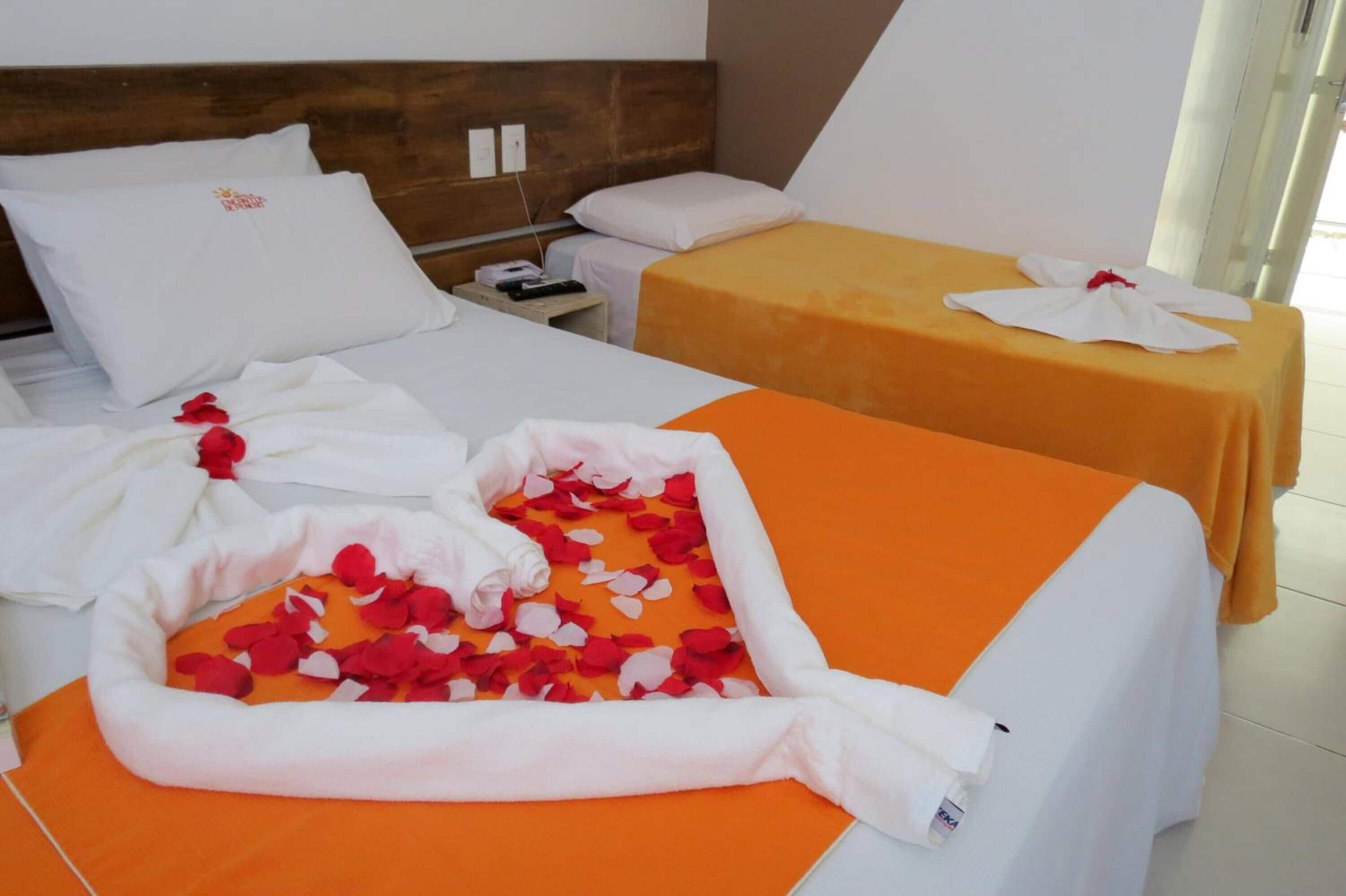 Une chambre d’hôtel avec deux lits, l’un recouvert d’une couvre-lit orange et l’autre d’une couvre-lit blanche. Des pétales de rose sont éparpillés sur le sol et une serviette blanche est posée sur le lit.