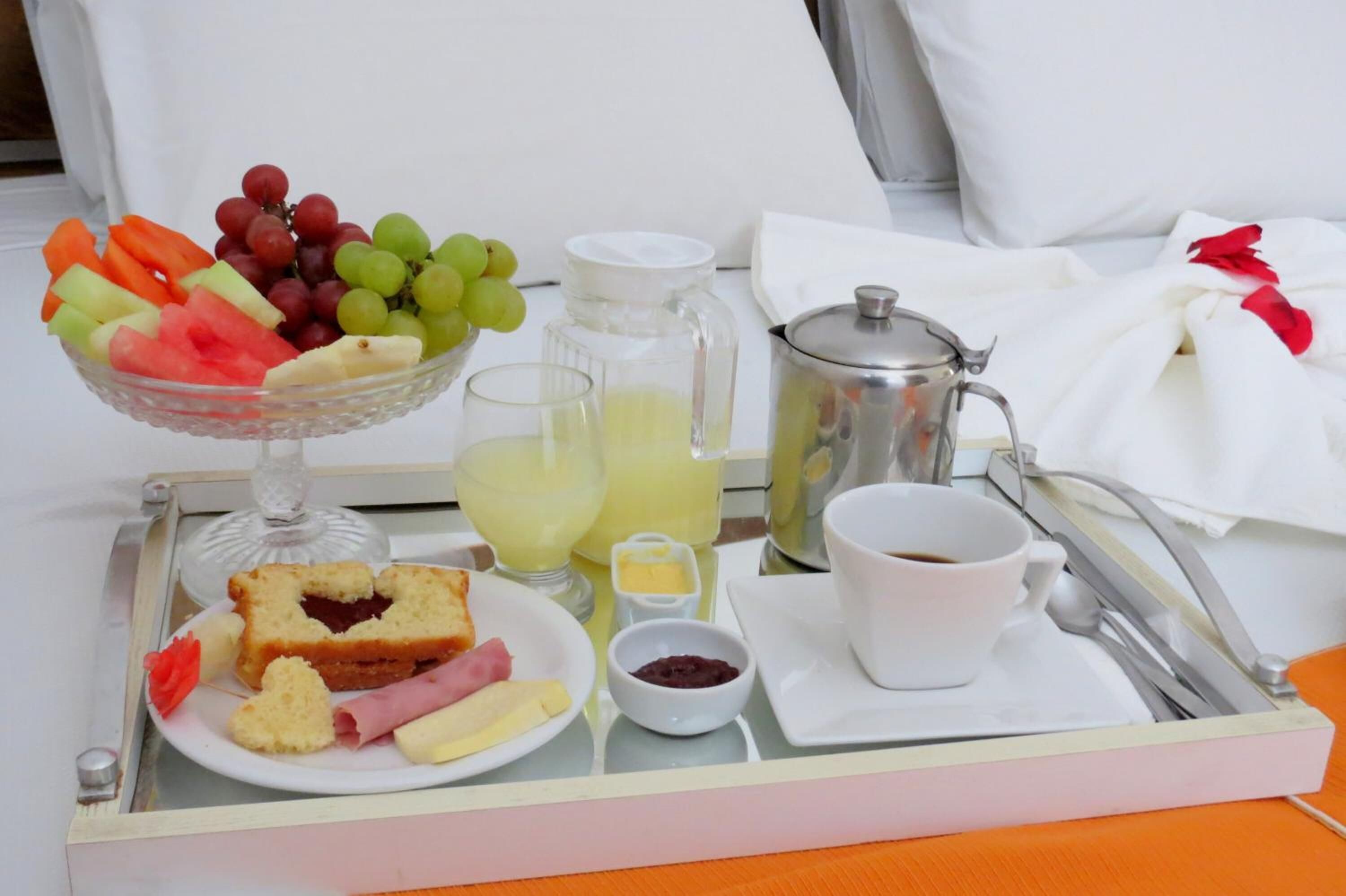 Una bandeja de desayuno con un bol de fruta, frutas cortadas, un sándwich de mermelada, una taza de café, una jarra de jugo y una cafetera.