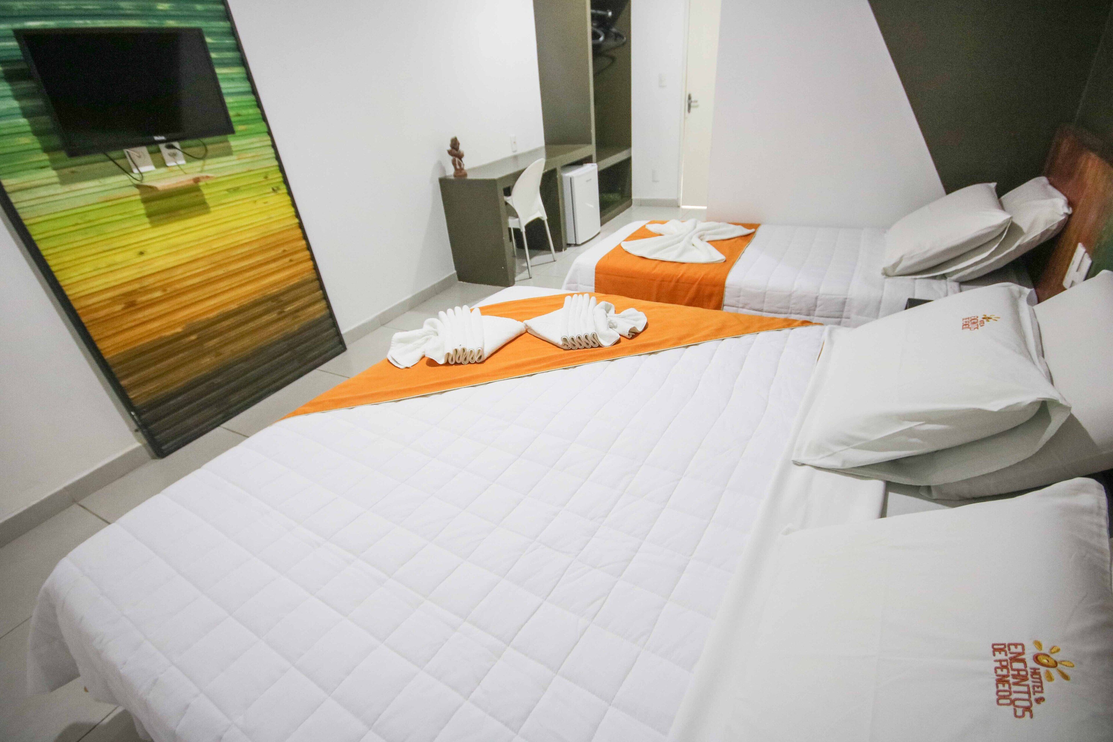 A hotel room with two beds, each with white bedding and orange blankets. There is a flat-screen TV mounted on the wall, a desk with a chair, and a mirror.