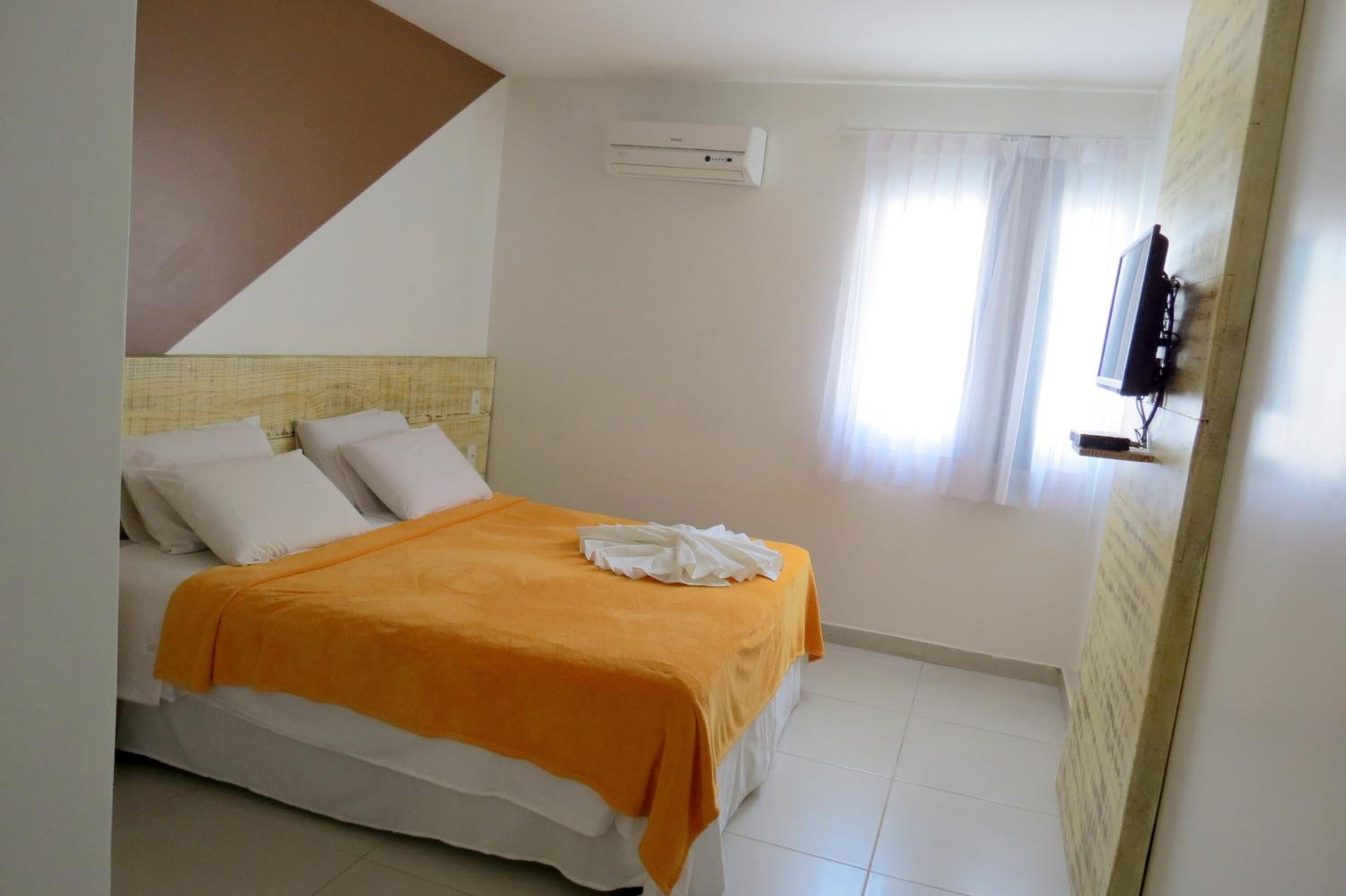 A bedroom with a bed, white pillows, an orange blanket, a TV mounted on the wall, and a window with curtains.