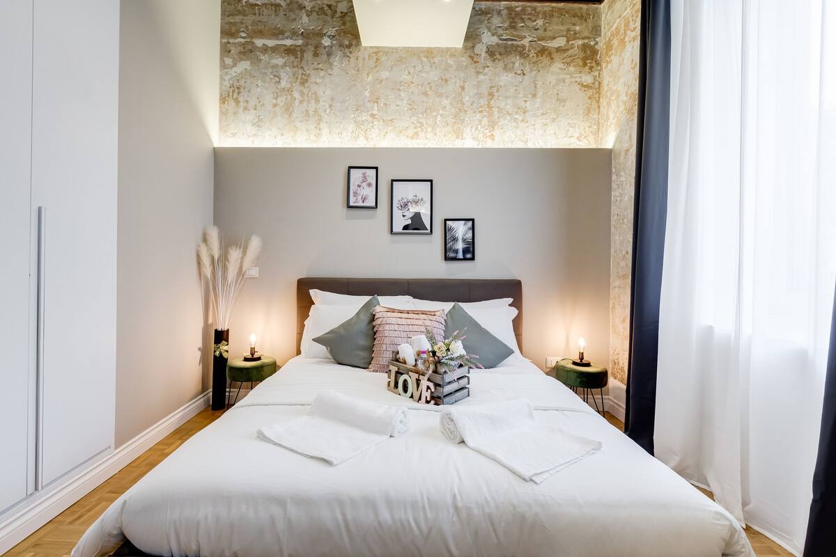 Una cama bien hecha con sábanas blancas, un cabecero con almohadones decorativos y una mesita de noche con una lámpara y un pequeño florero con flores.