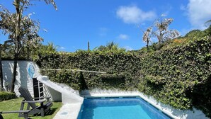 Una piscina con bordo piastrellato, circondata da un siepe e alcune poltrone da relax.