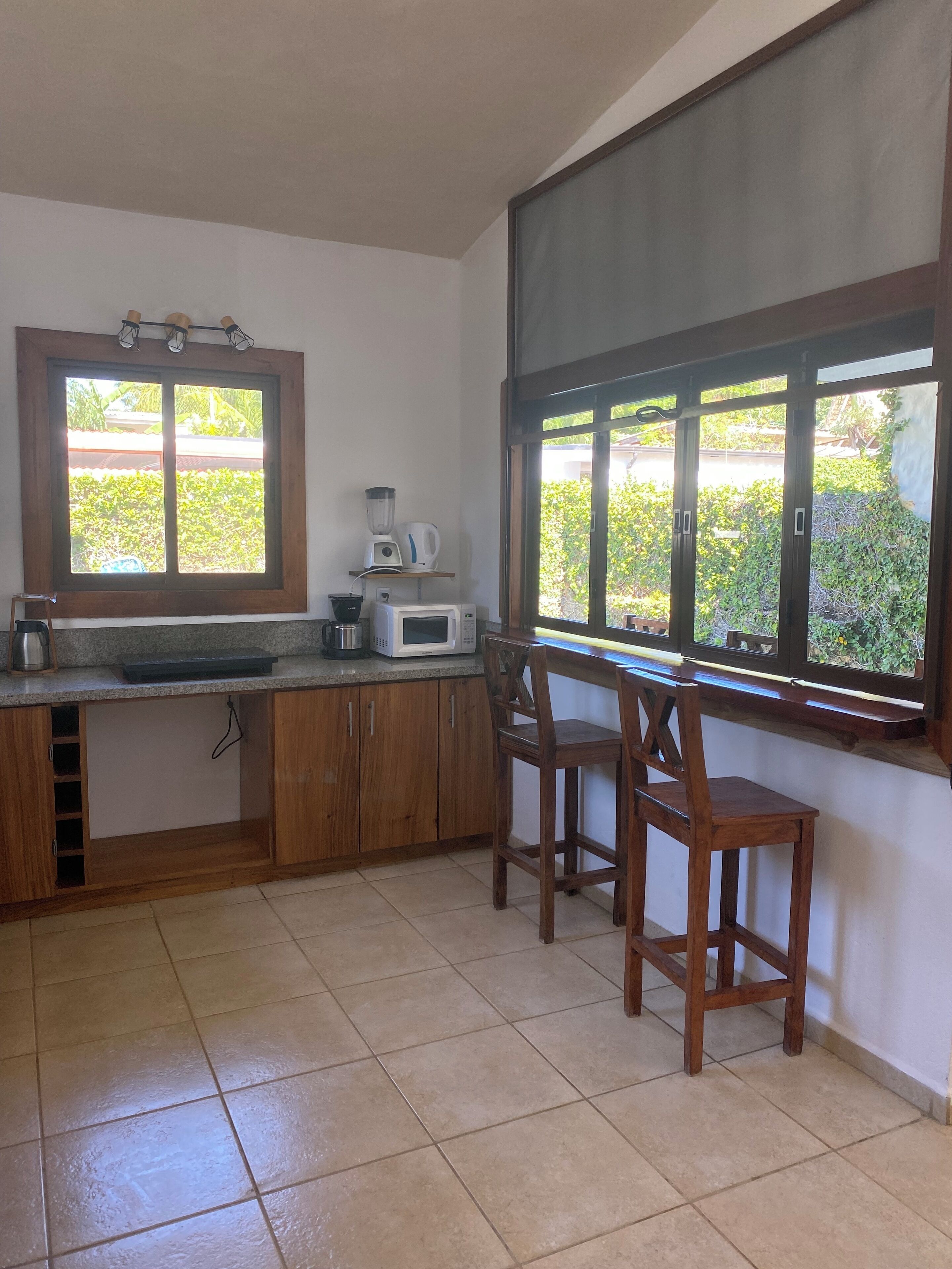 A kitchen with wooden cabinets, a countertop, a sink, a window with a view of greenery, and a chair.